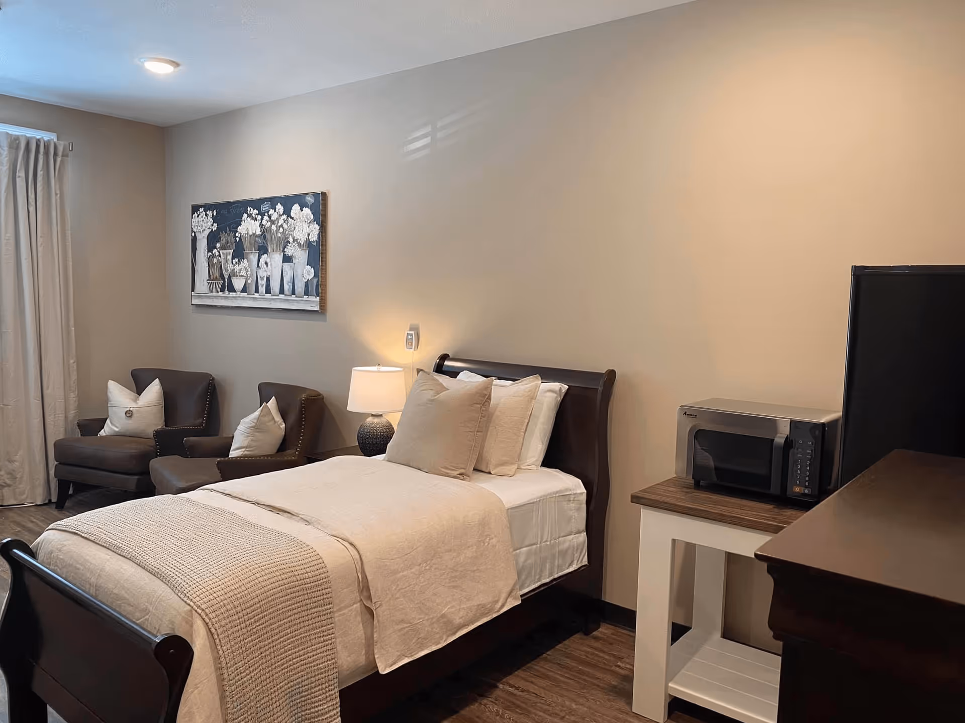 A cozy bedroom in Bellevue Assisted Living featuring a single bed with beige bedding and multiple pillows, two brown armchairs with white cushions near a window with beige curtains, a table lamp on a nightstand, a microwave on a small table, and a dark wooden dresser. A floral painting hangs on the beige wall above the armchairs.
