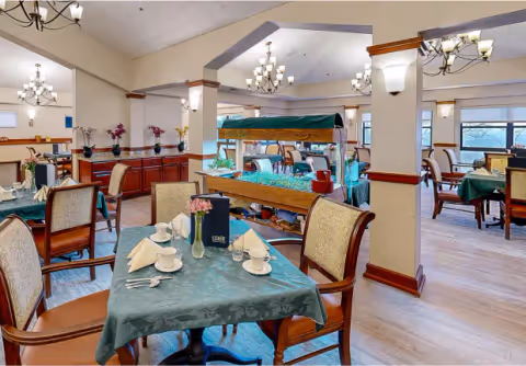 Bright dining room with set tables covered in green tablecloths, chandeliers, wooden chairs, and a central buffet station.