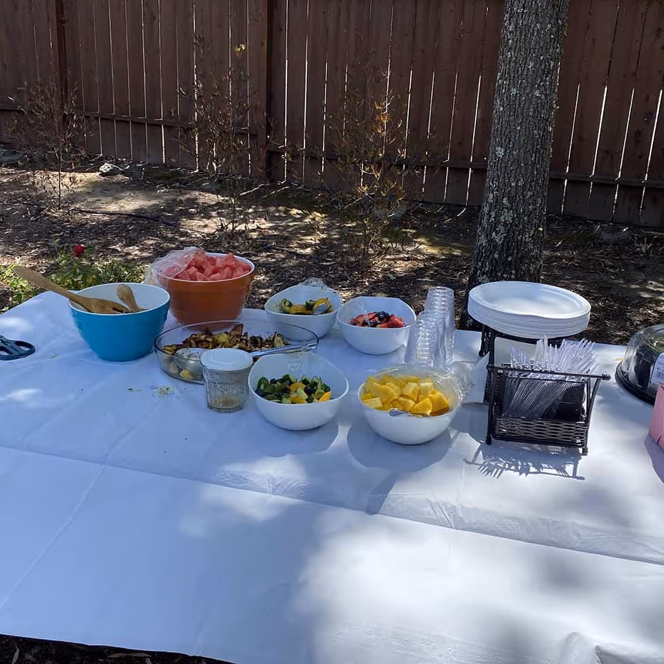 Outdoor table covered with a white tablecloth displaying bowls of various fruits and salads, plastic cups, plates, and utensils, set up near a wooden fence and trees.