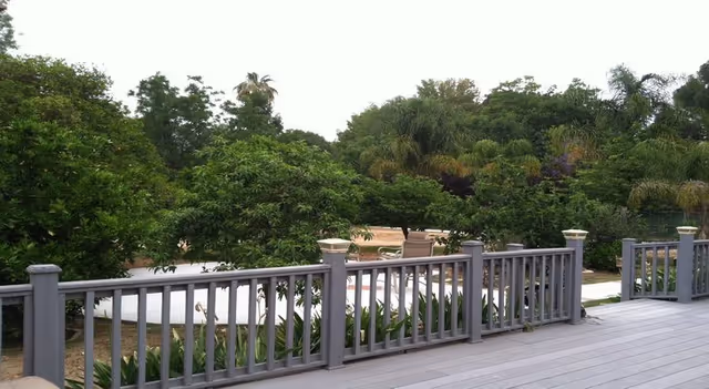 View from a wooden deck with gray railings overlooking a garden area with various green trees and plants under a bright sky.