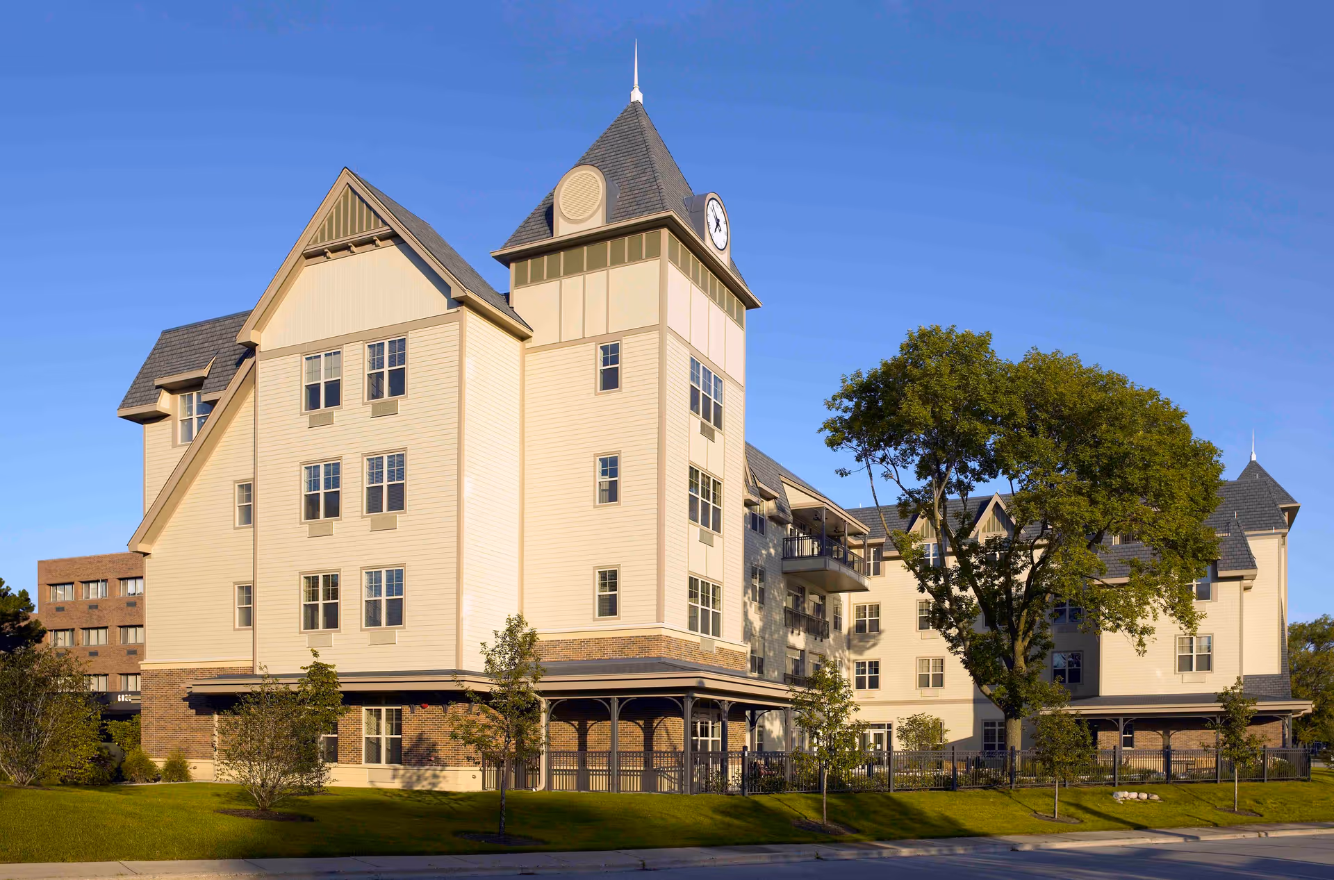 Exterior view of a multi-story senior living facility building with a clock tower, beige siding, and a sloped roof. The building is surrounded by green grass, trees, and a clear blue sky.
