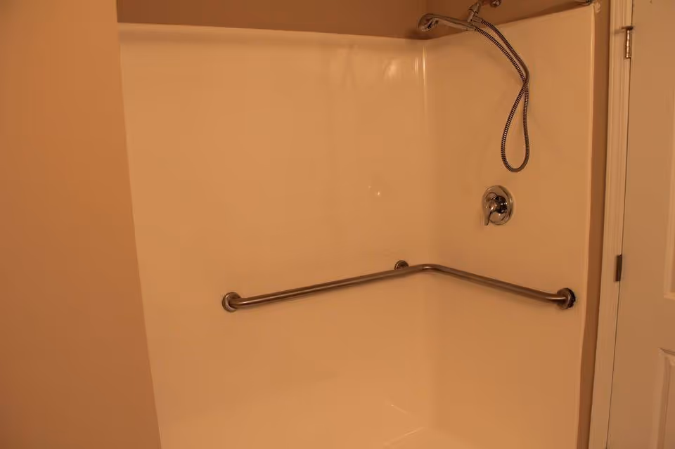 A shower area with beige walls, a white shower surround, a metal grab bar installed horizontally along the wall, a handheld showerhead attached to a flexible hose, and a single lever control valve. A white door is partially visible on the right side.