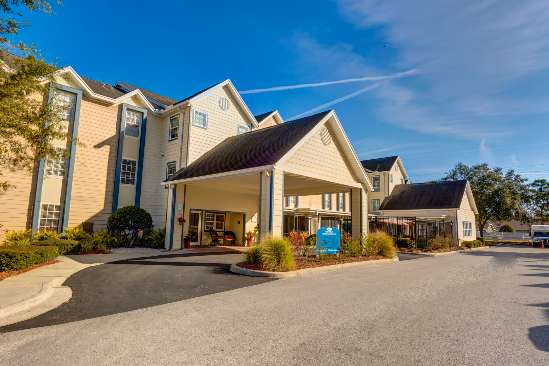 Exterior view of a senior living facility building with a covered entrance driveway, beige siding, multiple windows, and a clear blue sky. There are landscaped bushes and plants around the entrance area and a blue sign near the driveway.