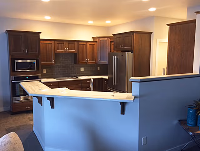 Interior view of a kitchen area with wooden cabinets, a stainless steel refrigerator, built-in microwave and oven, a light-colored countertop with a breakfast bar, and recessed ceiling lights.