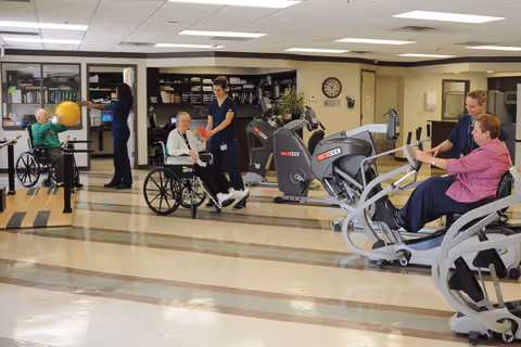 A physical therapy or rehabilitation room in a senior care facility with elderly individuals using exercise equipment and being assisted by caregivers. One person in a wheelchair is holding a red ball with a caregiver, another person in a wheelchair is holding a large yellow exercise ball, and two women are using seated rowing machines. The room has bright lighting and exercise equipment in the background.