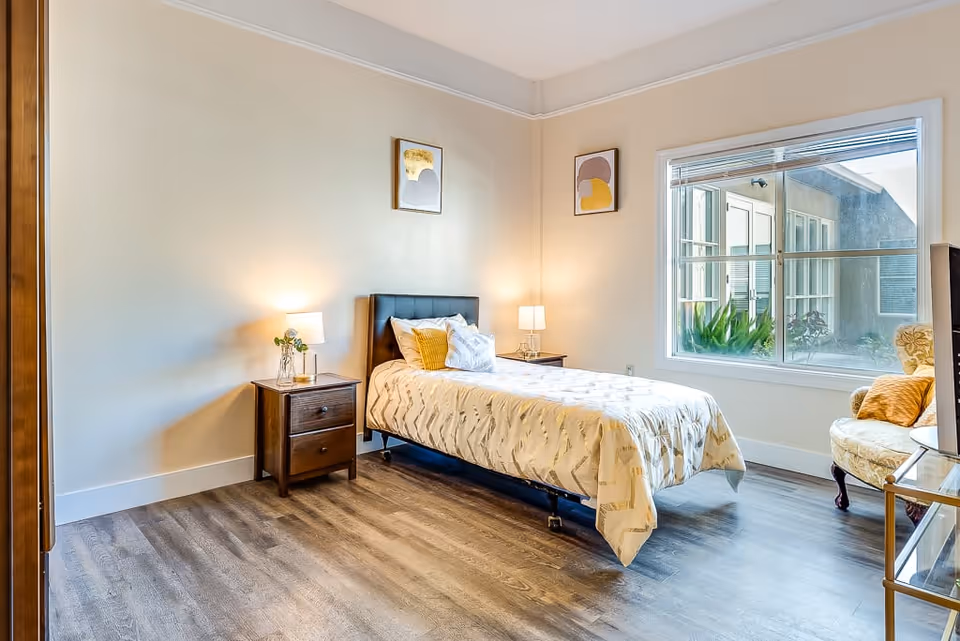 A bright and clean bedroom in a senior living facility featuring a single bed with a patterned bedspread and multiple pillows. There are two wooden nightstands on either side of the bed, each with a lamp. The room has light-colored walls with two framed abstract artworks hanging above the bed. A large window lets in natural light and shows some greenery outside. There is also a cushioned armchair with a pillow near the window and a TV on a stand in the corner.