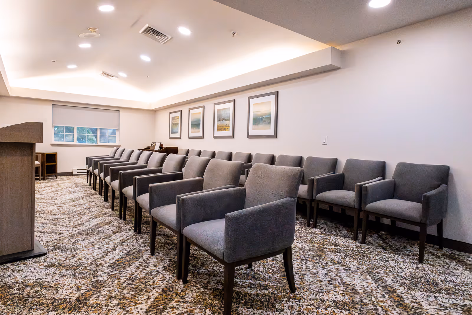 Rows of gray upholstered armchairs arranged in a carpeted meeting room with a podium and framed artwork on the wall.