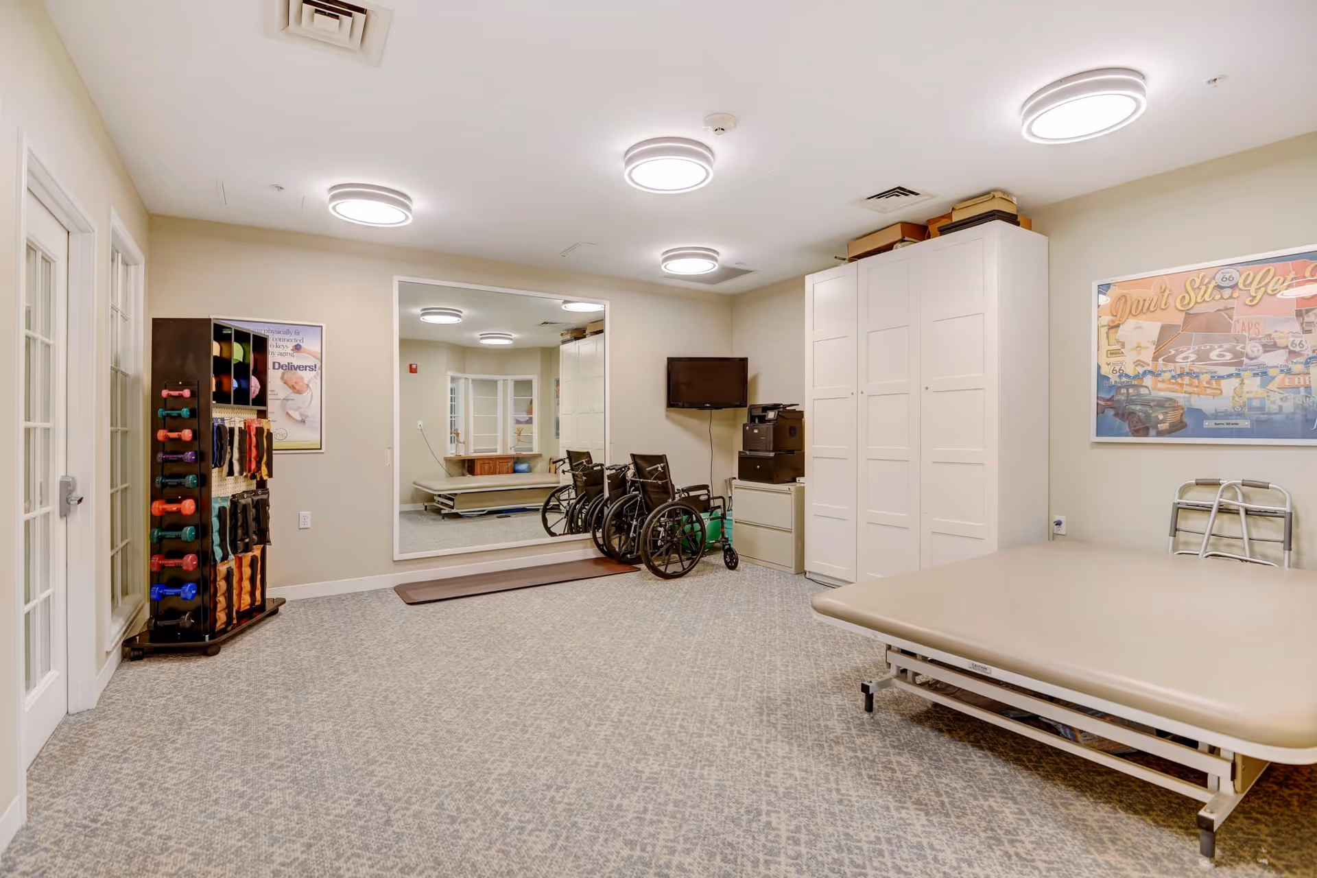 A physical therapy or rehabilitation room with exercise equipment including colorful dumbbells on a rack, two wheelchairs, a walker, a large mirror on the wall, a therapy table, and a TV mounted on the wall. The room has beige walls, patterned carpet, and bright circular ceiling lights.