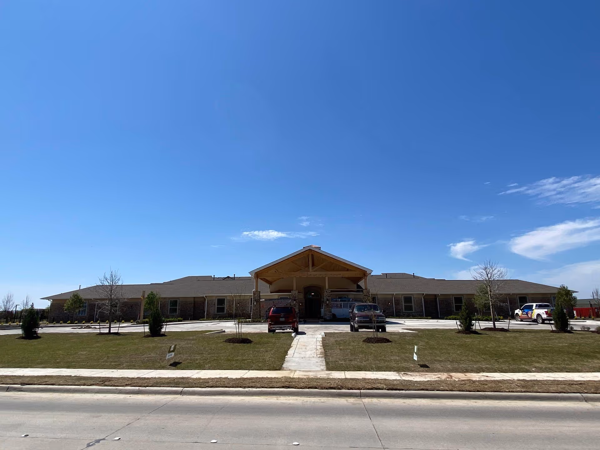 Single-story memory care building with a covered entrance, parked vehicles, and a grassy front lawn under a clear blue sky.