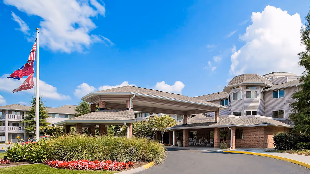 Exterior view of Holiday Echo Ridge facility showing a covered entrance with a driveway, landscaped garden with flowers and bushes, a flagpole with three flags, and a multi-story building under a blue sky with some clouds.