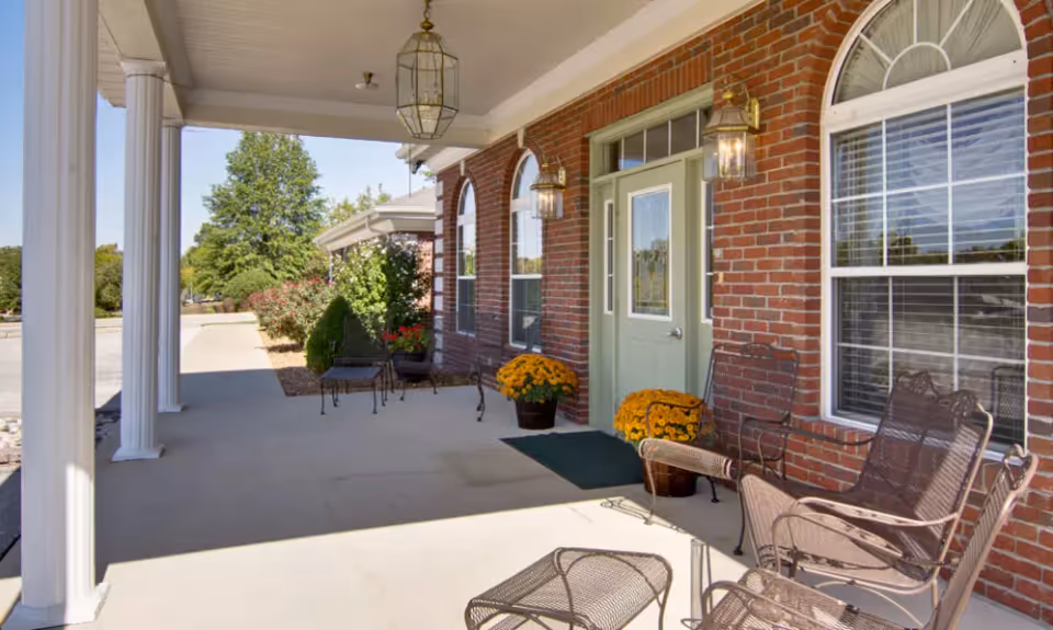 Covered outdoor patio area with white columns, brick wall, green door, windows with white blinds, metal chairs and tables, potted yellow flowers, and hanging lantern-style lights.