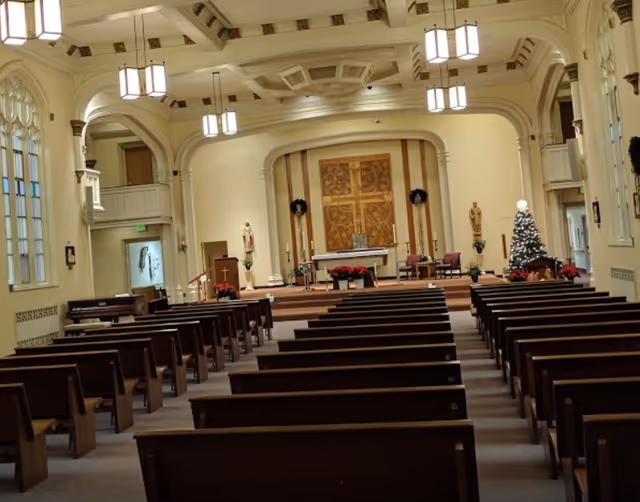 Interior view of a chapel sanctuary with rows of wooden pews leading to an altar decorated for Christmas with a tree and wreaths.
