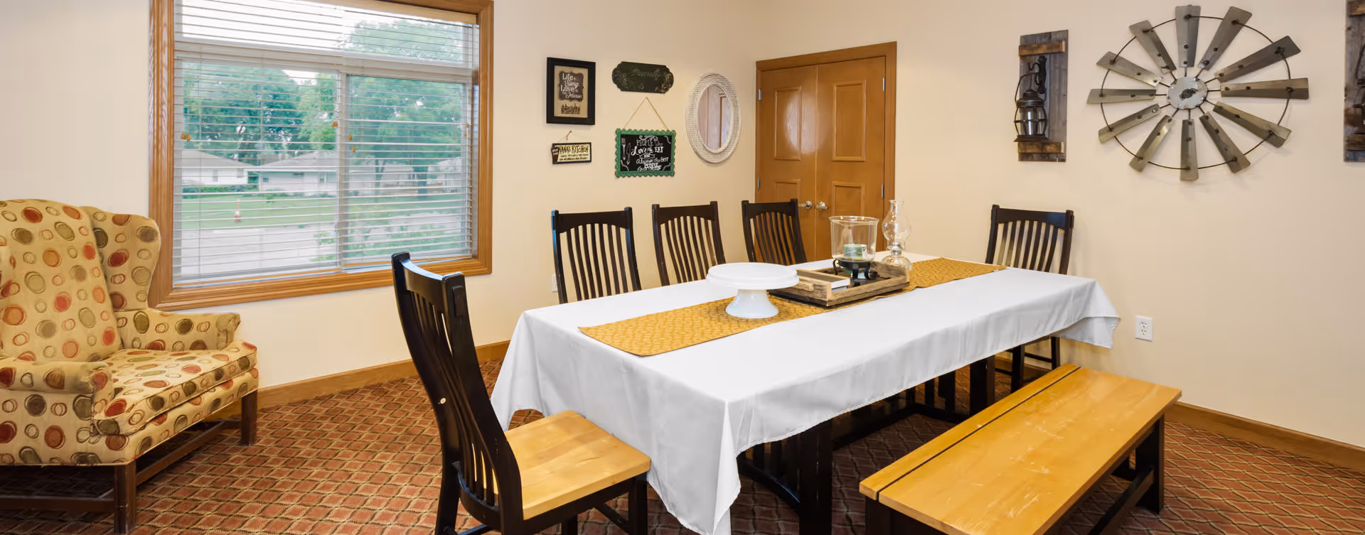 A dining room with a rectangular table covered with a white tablecloth and a yellow table runner. The table is surrounded by six wooden chairs and a wooden bench. On the table are decorative items including a tray, a glass candle holder, and a cake stand. To the left is a patterned armchair near a window with blinds, and the walls are decorated with various wall hangings including a large clock and framed signs.