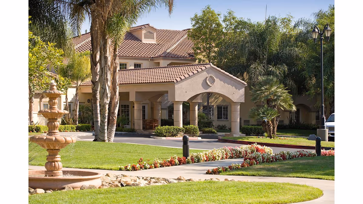 Exterior view of a senior living facility named Park Terrace featuring a beige stucco building with a red tile roof, a covered entrance supported by columns, well-maintained green lawns, palm trees, a flower bed with red and white flowers, a stone fountain, and a black street lamp.