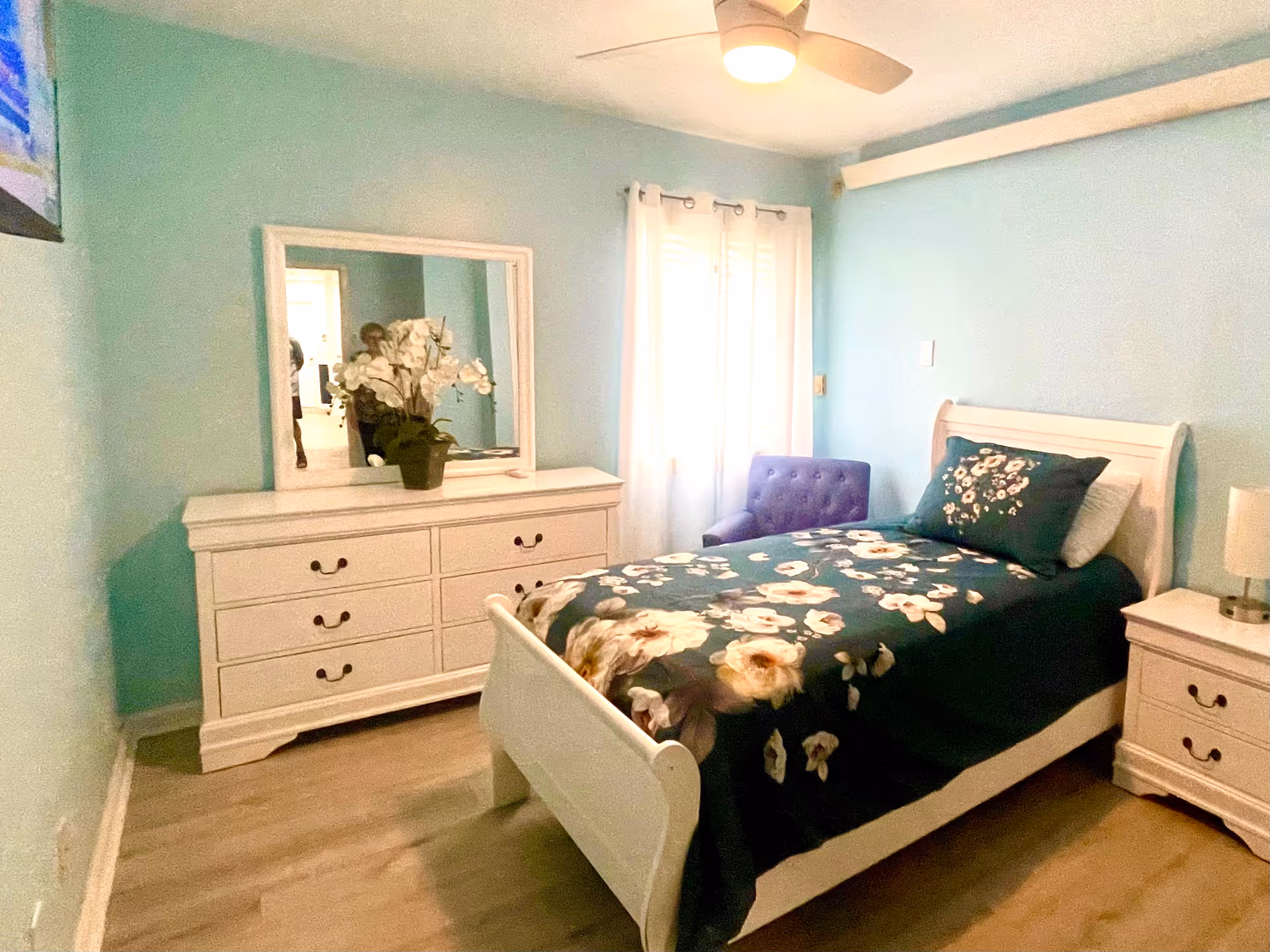 A cozy bedroom with light blue walls featuring a white wooden bed with floral bedding, a matching white dresser with a mirror and a potted plant, a white nightstand with a lamp, a purple upholstered chair near a window with white curtains, and a ceiling fan with a light.