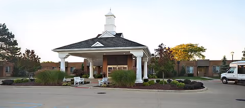 Front exterior view of Rivergate Health Care Center featuring a covered entrance with white columns, a small cupola on the roof, landscaped bushes, benches, and a driveway with a parked white van on the right side.