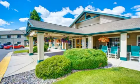 Exterior view of a senior living facility building with a covered entrance, green bushes, and a well-maintained lawn under a bright blue sky with scattered clouds. There are chairs on the porch and a parking area with cars visible in the background.