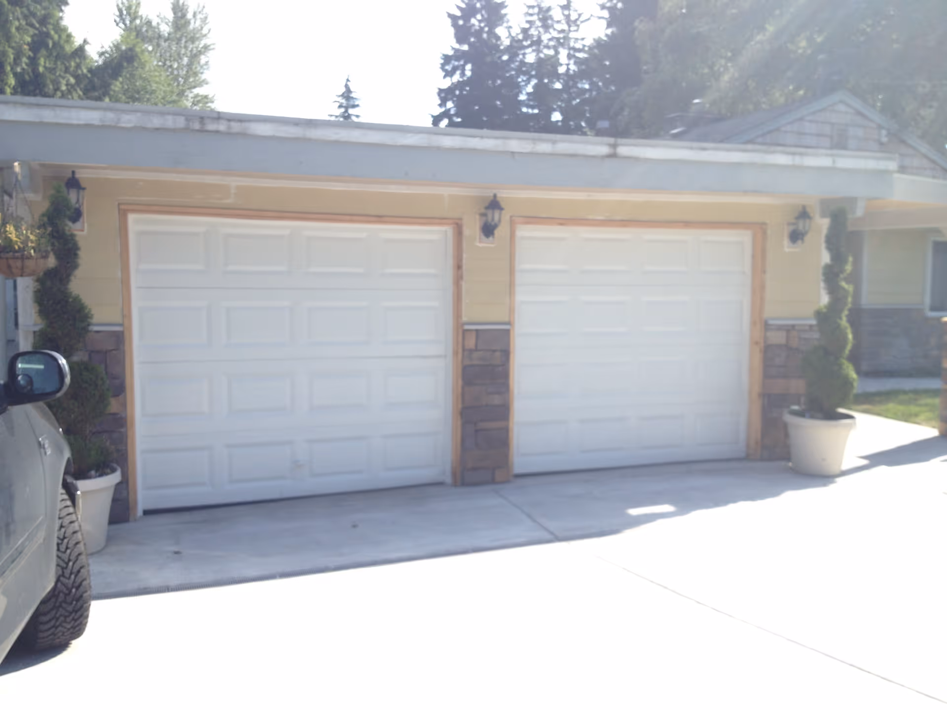 Exterior view of a building with two white garage doors side by side, flanked by potted topiary plants. A vehicle is partially visible on the left side of the image.