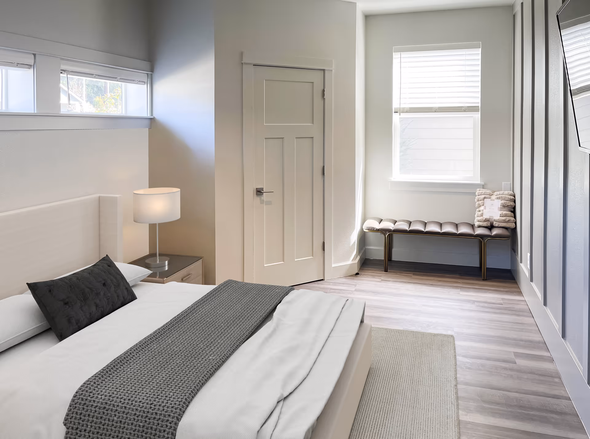 A bright and modern bedroom with a neatly made bed featuring white linens and a gray knitted throw. There is a bedside table with a lamp, a closed white door, a window with blinds, and a cushioned bench with folded blankets on it. The room has light-colored walls and wood flooring.
