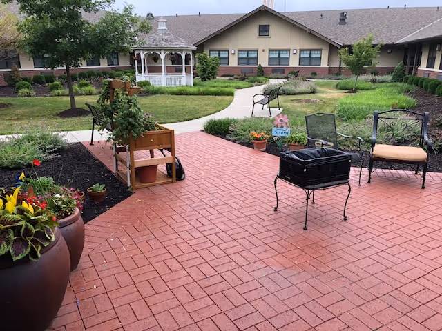 Outdoor patio area with red brick flooring, metal chairs with cushions, a small black grill, potted plants, and a wooden planter. In the background, there is a white gazebo, green grass, shrubs, and a building with beige walls and multiple windows.