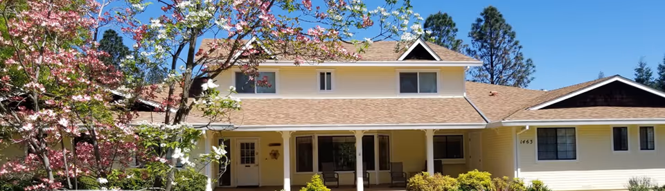 Front exterior view of a two-story yellow house with a brown roof, a covered porch with chairs, surrounded by green bushes and trees with pink and white blossoms under a clear blue sky.