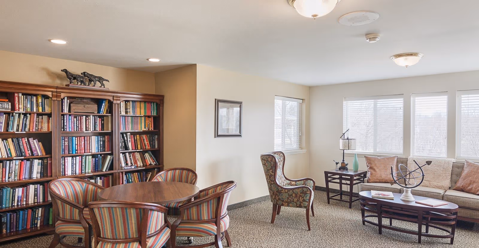 A cozy living room area with a round wooden table surrounded by four striped upholstered chairs. Behind the table is a large wooden bookshelf filled with books and a decorative sculpture on top. To the right, there is a patterned armchair, a side table with a lamp and decorative items, and a beige sofa with several pillows. The room has multiple windows letting in natural light and neutral-colored walls and carpet.