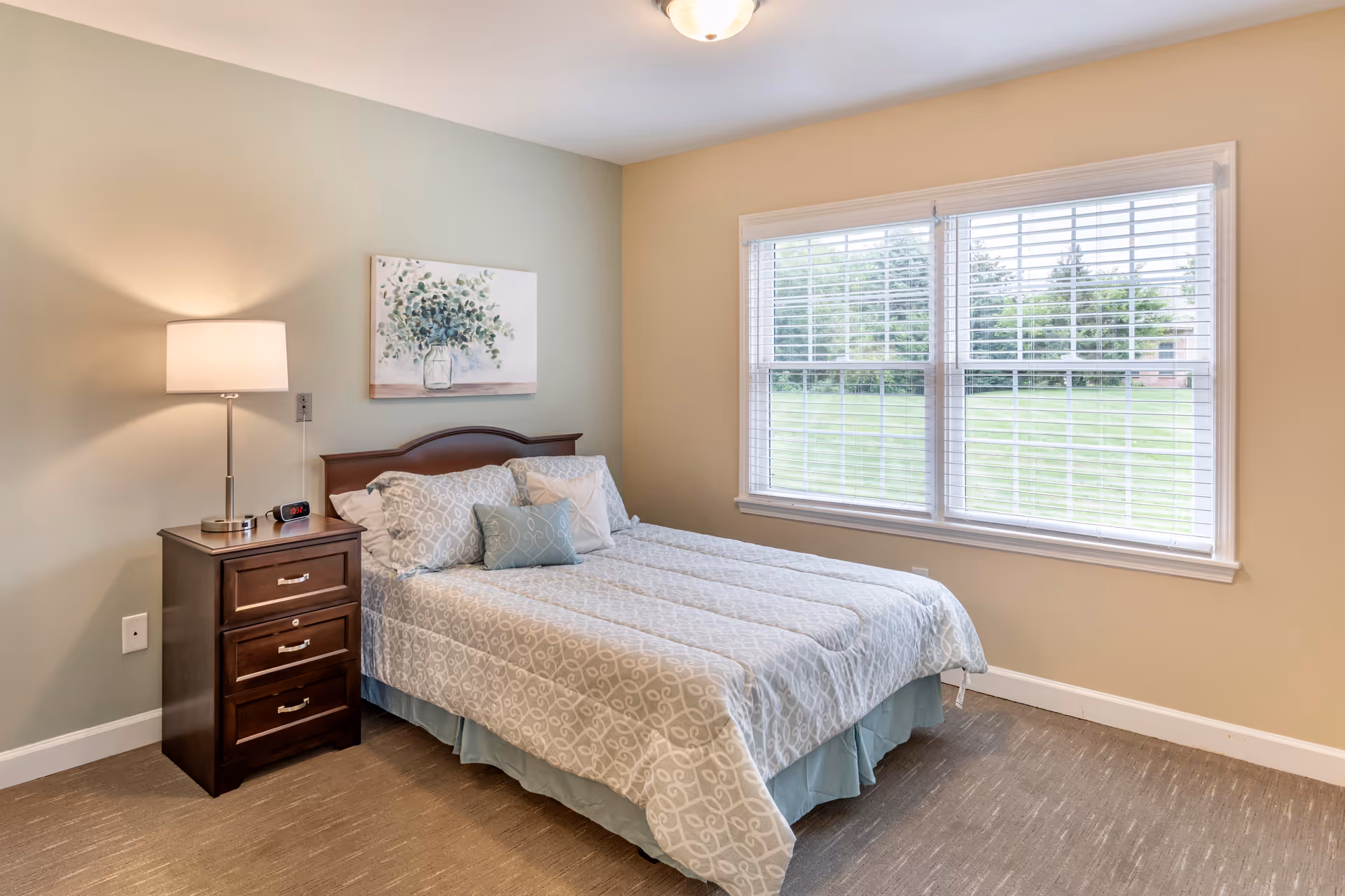 A neatly made bed with patterned bedding and multiple pillows in a bedroom. Next to the bed is a wooden nightstand with three drawers, a lamp, and a digital alarm clock. A painting of a vase with greenery hangs on the wall above the bed. Large windows with white blinds let in natural light and show a green lawn outside.