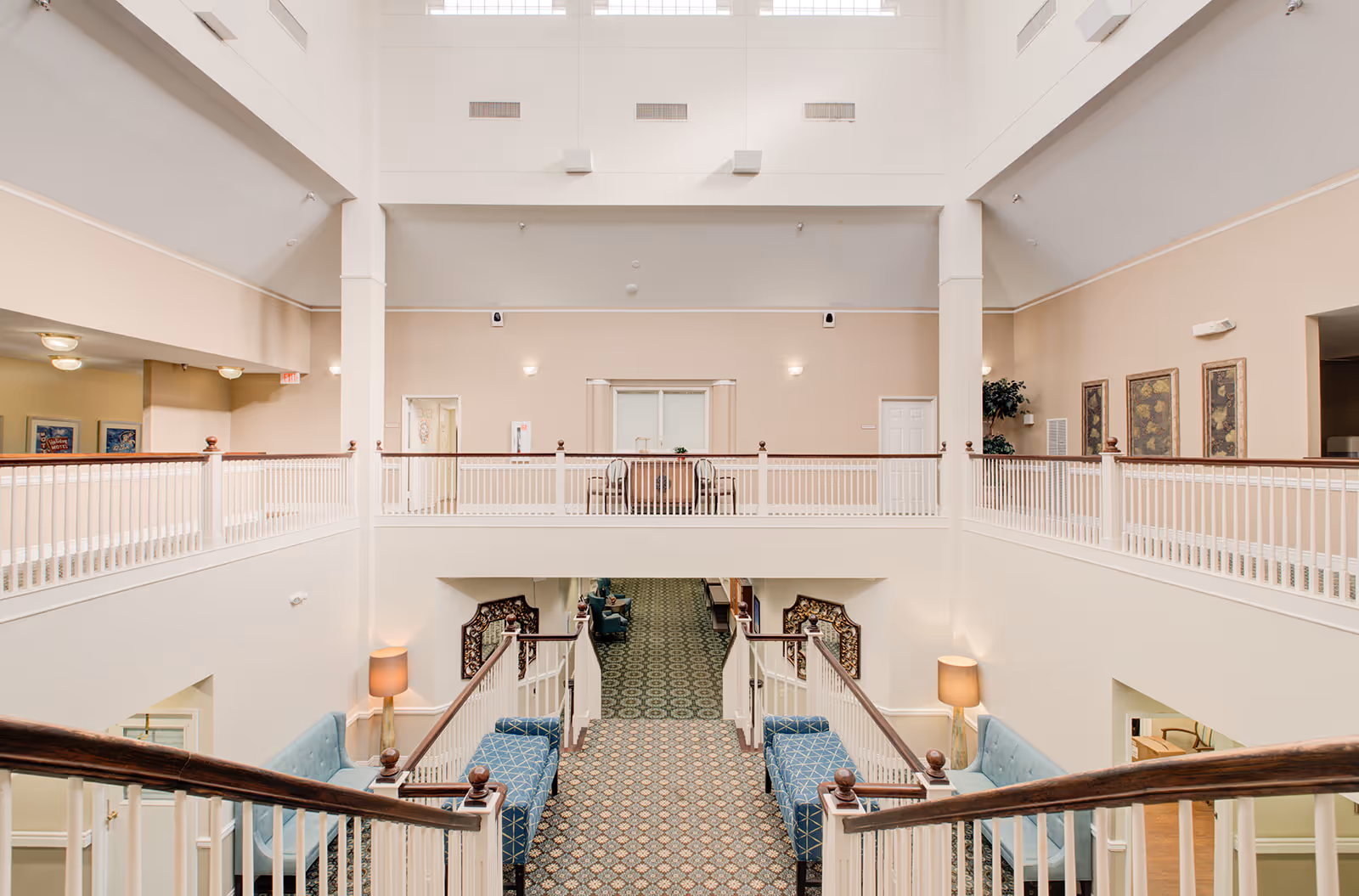 Interior view of a senior living facility with a wide staircase leading down to a hallway. The area features beige walls, white railings, patterned carpet, blue upholstered chairs, and decorative wall art. The space is well-lit with ceiling lights and lamps.