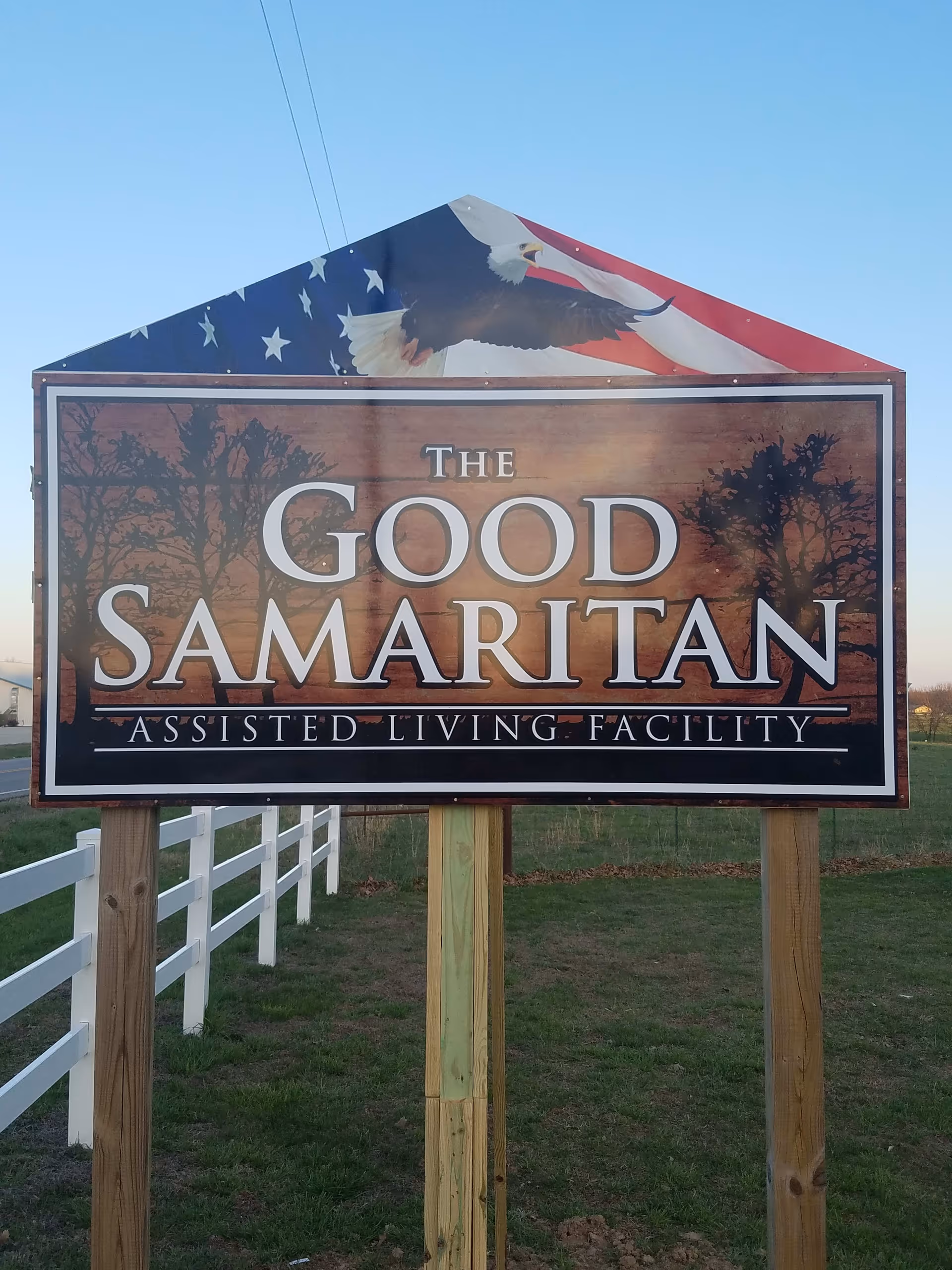 A large outdoor sign for The Good Samaritan Assisted Living Facility featuring an American flag and a bald eagle at the top, mounted on wooden posts with a white fence and grassy area in the background.