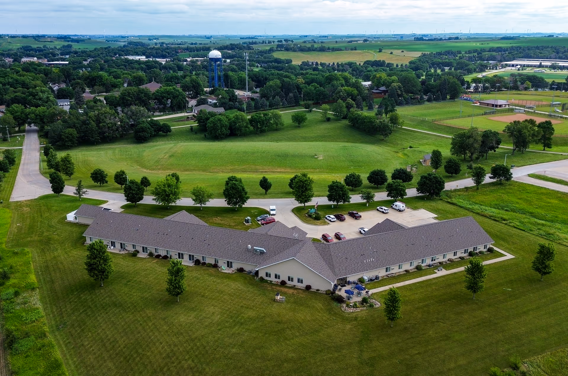 Aerial view of a single-story senior living facility building surrounded by lawns, trees, a parking lot, and nearby fields.