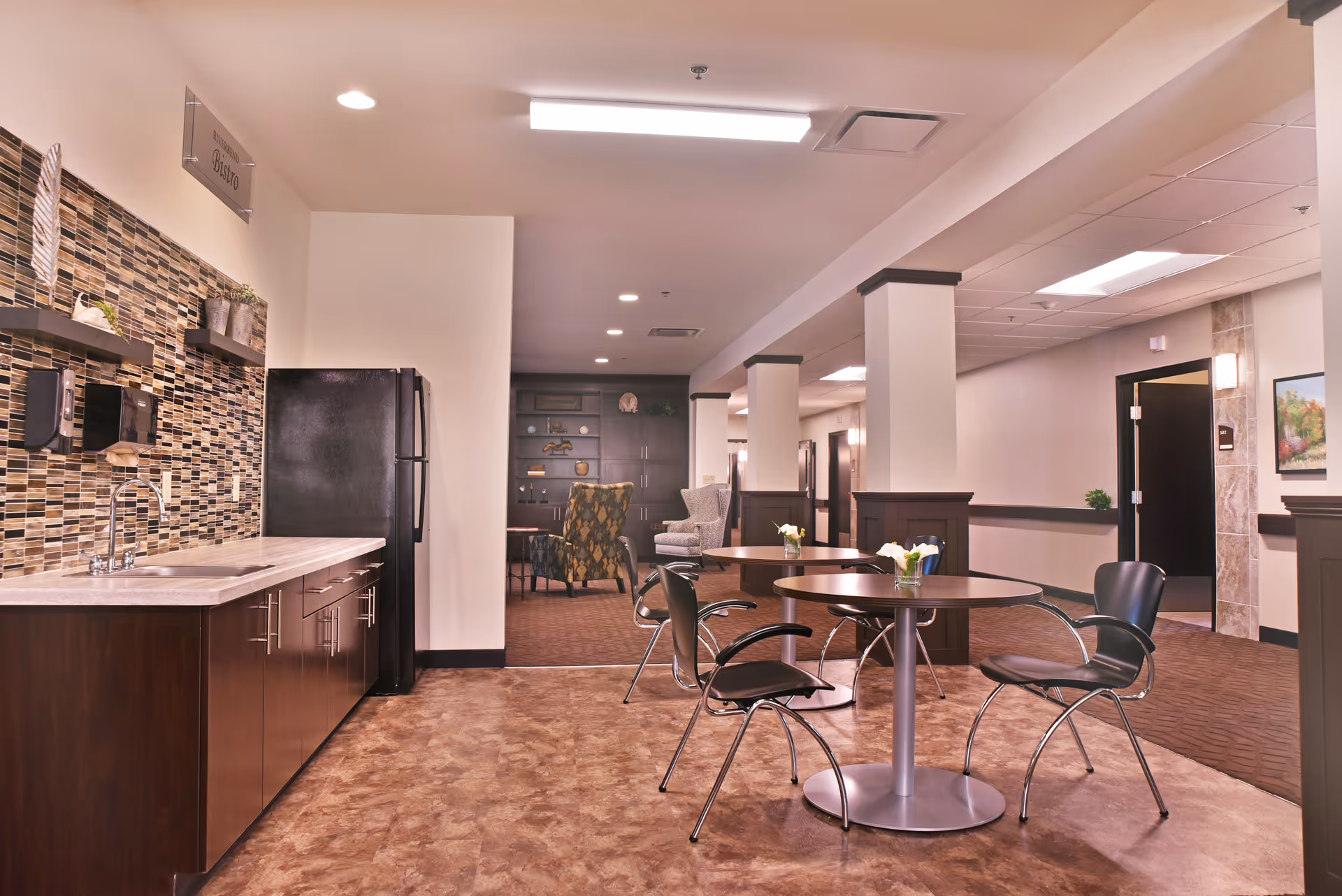 Interior view of a senior living facility common area with a small kitchenette on the left featuring a sink, microwave, and refrigerator. In the foreground, there are round tables with black chairs and small flower arrangements. The background shows a seating area with armchairs and built-in shelves.