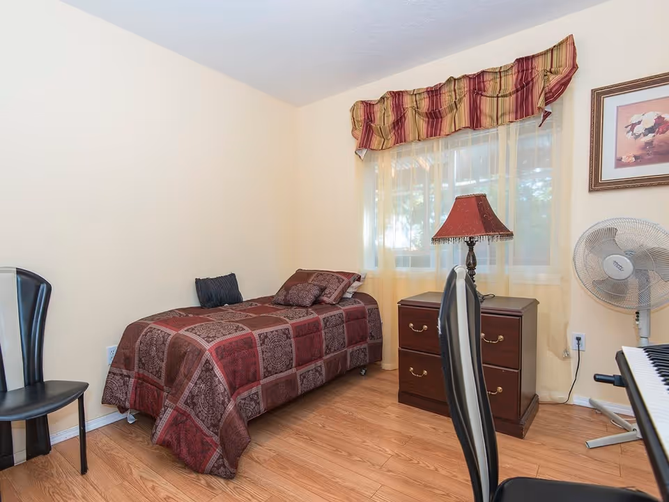 A small bedroom with a single bed covered in a red and brown patterned bedspread. There is a black chair next to the bed, a wooden nightstand with a red lampshade, a standing fan, and a window with sheer curtains and a striped valance. The floor is wooden, and there is a framed picture on the wall.