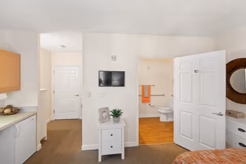 Studio-style bedroom with a small kitchenette on the left, a dresser and wall-mounted TV in the center, and an open door revealing a bathroom.