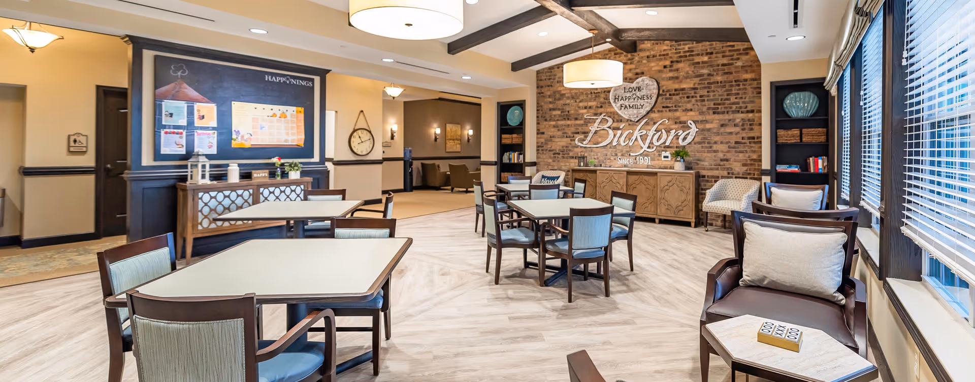 A bright and spacious common area in a senior living facility with several tables and chairs arranged for socializing or activities. The room features a brick accent wall with the words 'Love Happiness Family Bickford Since 1991' displayed. There are large windows with blinds on the right side, a bulletin board labeled 'Happenings' on the left wall, and comfortable seating areas with armchairs and small tables.