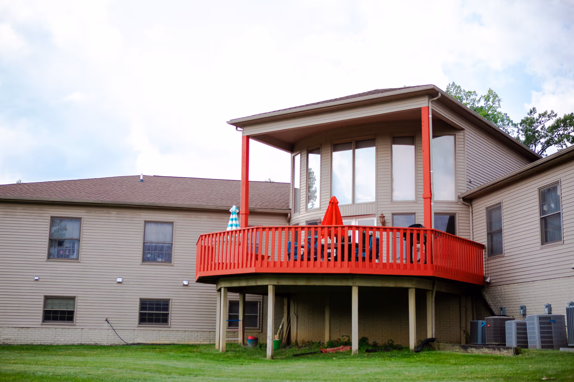 Exterior view of a senior living facility building with beige siding and a brown roof. The building features a raised wooden deck painted bright red with a red umbrella and outdoor furniture. The deck is supported by wooden posts and overlooks a grassy lawn. Several windows are visible on the building, and there are air conditioning units on the right side near the ground.