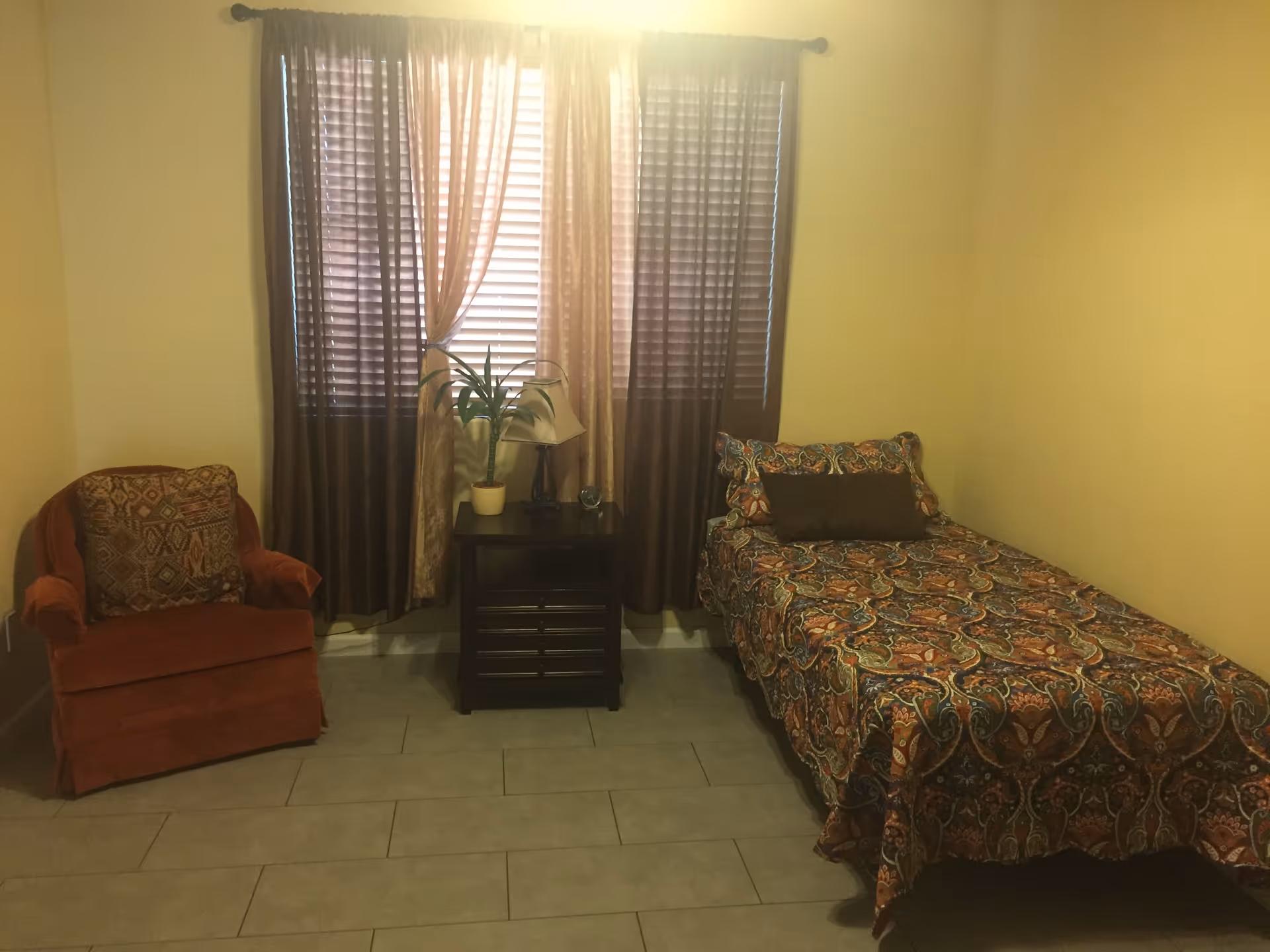 A small bedroom with a single bed covered in a colorful patterned bedspread and a matching pillow. Next to the bed is a dark wooden nightstand with a potted plant and a small lamp. To the left of the nightstand is an upholstered armchair with a patterned cushion. The room has beige walls and a window with brown and beige curtains letting in some light.
