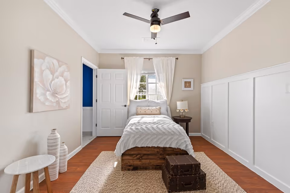 A bright and cozy bedroom with a single bed centered against the back wall, adorned with white bedding and pillows. A window with white curtains is behind the bed, allowing natural light to fill the room. To the right of the bed is a small round wooden nightstand with a lamp. The walls are painted beige with white wainscoting on the right side. On the left wall, there is a large floral painting and two decorative vases on the floor next to a small round white table. The floor is wooden with a beige area rug. A ceiling fan with a light is mounted on the white ceiling. An open door on the left side leads to another room painted blue.