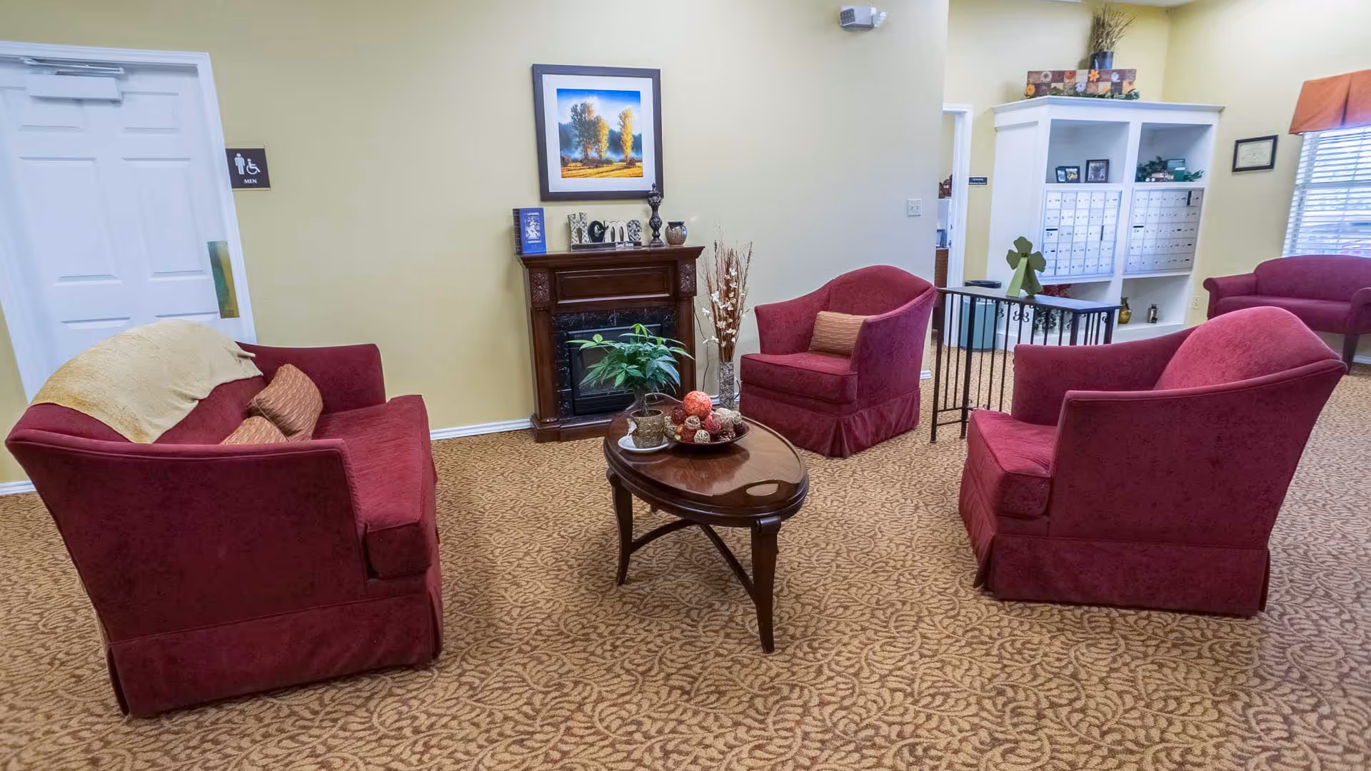 A cozy sitting area with red upholstered chairs and a sofa arranged around a wooden coffee table with decorative items. There is a small fireplace against a yellow wall with framed artwork above it. The carpet has a patterned design, and there is a door with a men's restroom sign on the left side. In the background, there are mailboxes and a window with blinds.