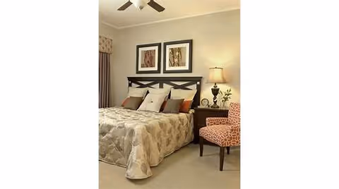 A neatly made bed with a beige patterned comforter and multiple pillows in a bedroom. There are two framed pictures hanging above the headboard, a bedside table with a lamp and a small plant, and an upholstered chair with a red and white pattern next to the bed.