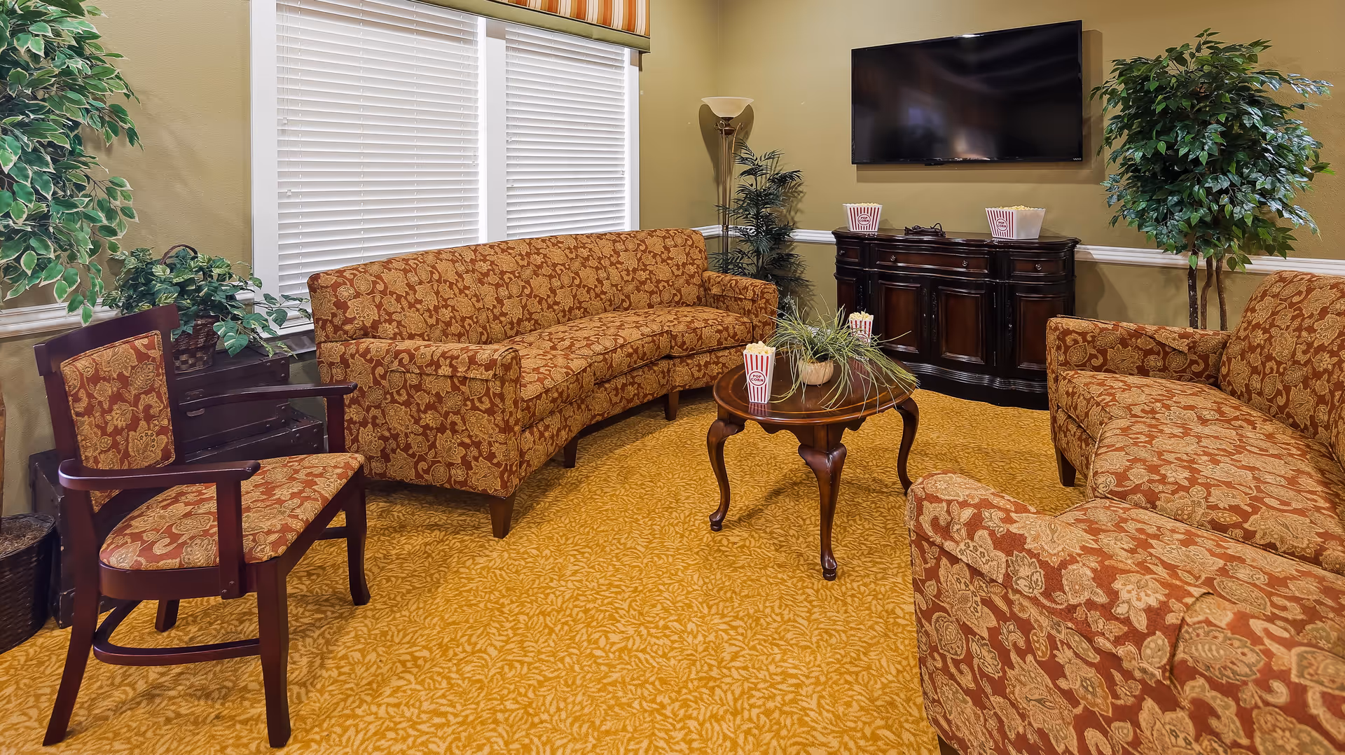 A cozy living room with patterned orange sofas and a matching armchair arranged around a wooden coffee table with a plant and popcorn containers. A flat-screen TV is mounted on the wall above a dark wooden cabinet. The room has beige walls, a yellow patterned carpet, two large windows with blinds and a striped valance, and several green potted plants.