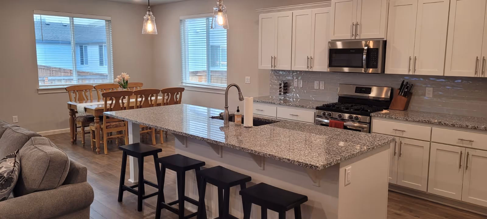 A modern kitchen and dining area with a large granite island countertop featuring a sink and three black stools. The kitchen has white cabinets, a stainless steel stove, microwave, and a knife block. In the background, there is a wooden dining table with six chairs and two windows letting in natural light. A gray sofa is partially visible on the left side.