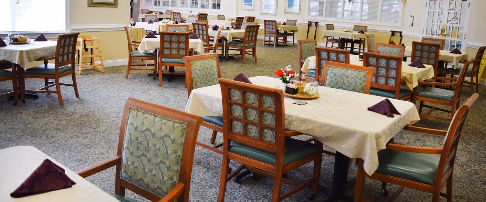 Bright dining room with multiple tables and wooden chairs arranged with tablecloths, napkins, and centerpieces.