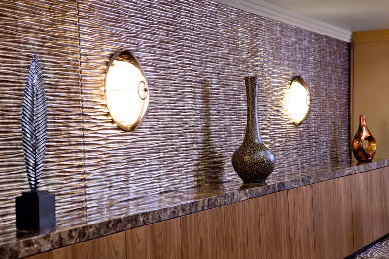 A decorative interior wall with a textured, wavy surface illuminated by two round wall sconces. Below the wall is a wooden ledge with a marble-like top, displaying three decorative vases of different shapes and colors.
