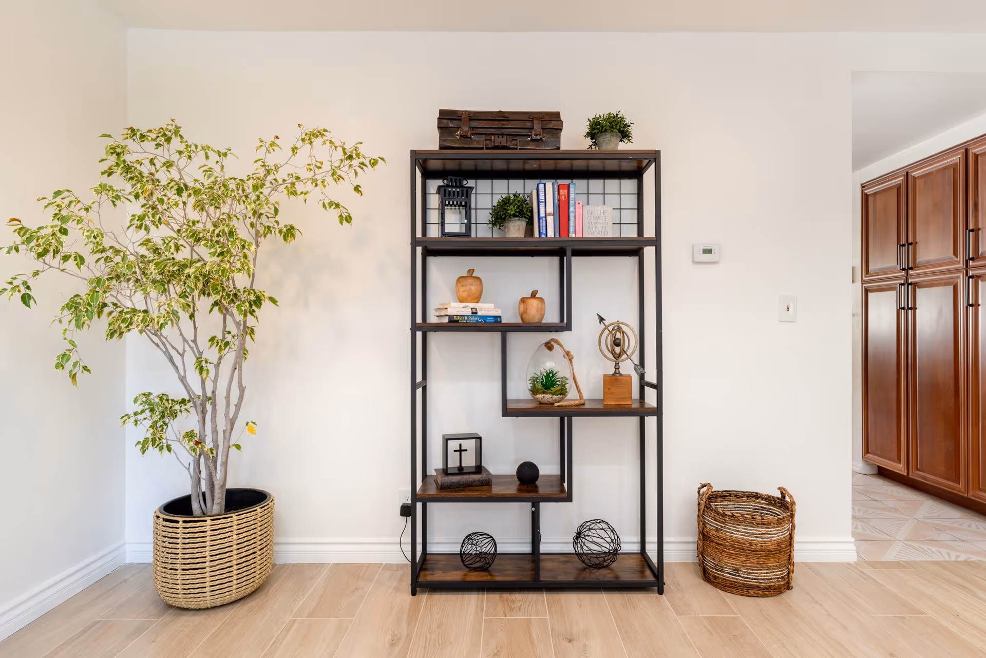 A decorative metal and wood shelving unit against a white wall, flanked by a tall potted plant on the left and a woven basket on the right.