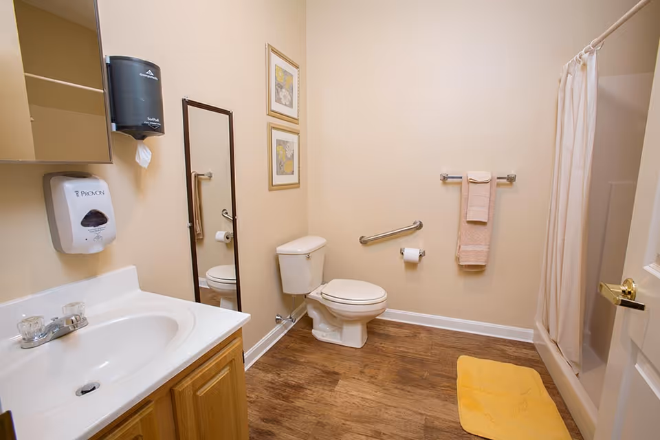 A clean bathroom with a white sink and wooden cabinet, a toilet, a shower with a white curtain, a yellow bath mat on the wooden floor, a towel rack with a beige towel, a wall-mounted soap dispenser, a paper towel dispenser, a full-length mirror, and two framed floral pictures on the wall.