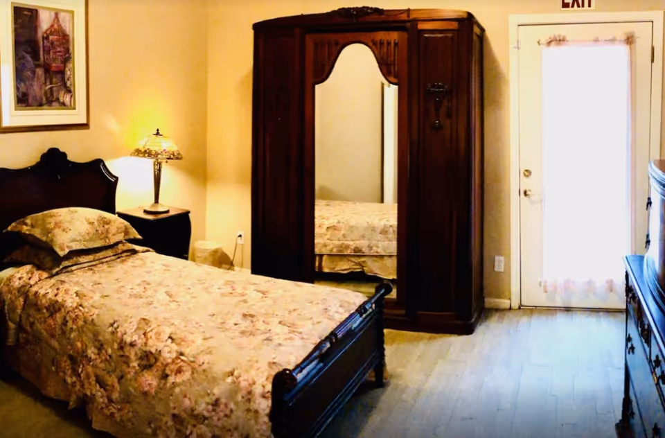 A cozy bedroom with a single bed in floral bedding, a wooden armoire with a mirrored door, a nightstand and lamp, and a door with an EXIT sign.