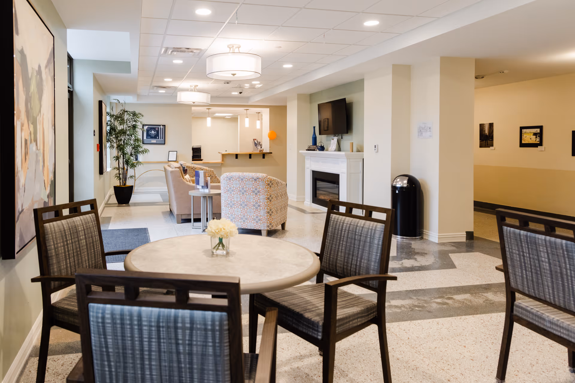 A bright and spacious common area in Bardwell Residences featuring a round table with four chairs in the foreground, a seating area with armchairs and a sofa near a fireplace with a mounted TV, modern ceiling lights, and artwork on the walls.
