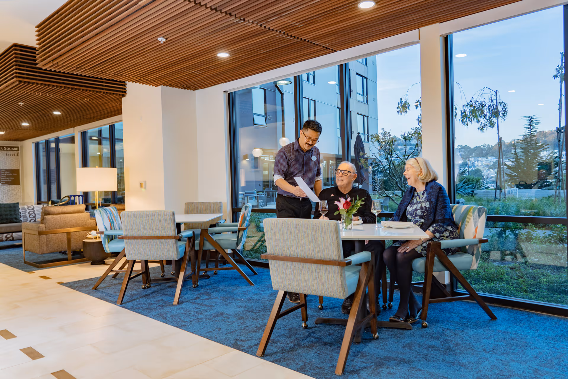 A dining area in an assisted living facility with large windows showing an outdoor view. Two elderly people are seated at a table with wine glasses and a flower vase, while a staff member stands next to them showing a paper. The room has modern furniture and a wooden slatted ceiling.