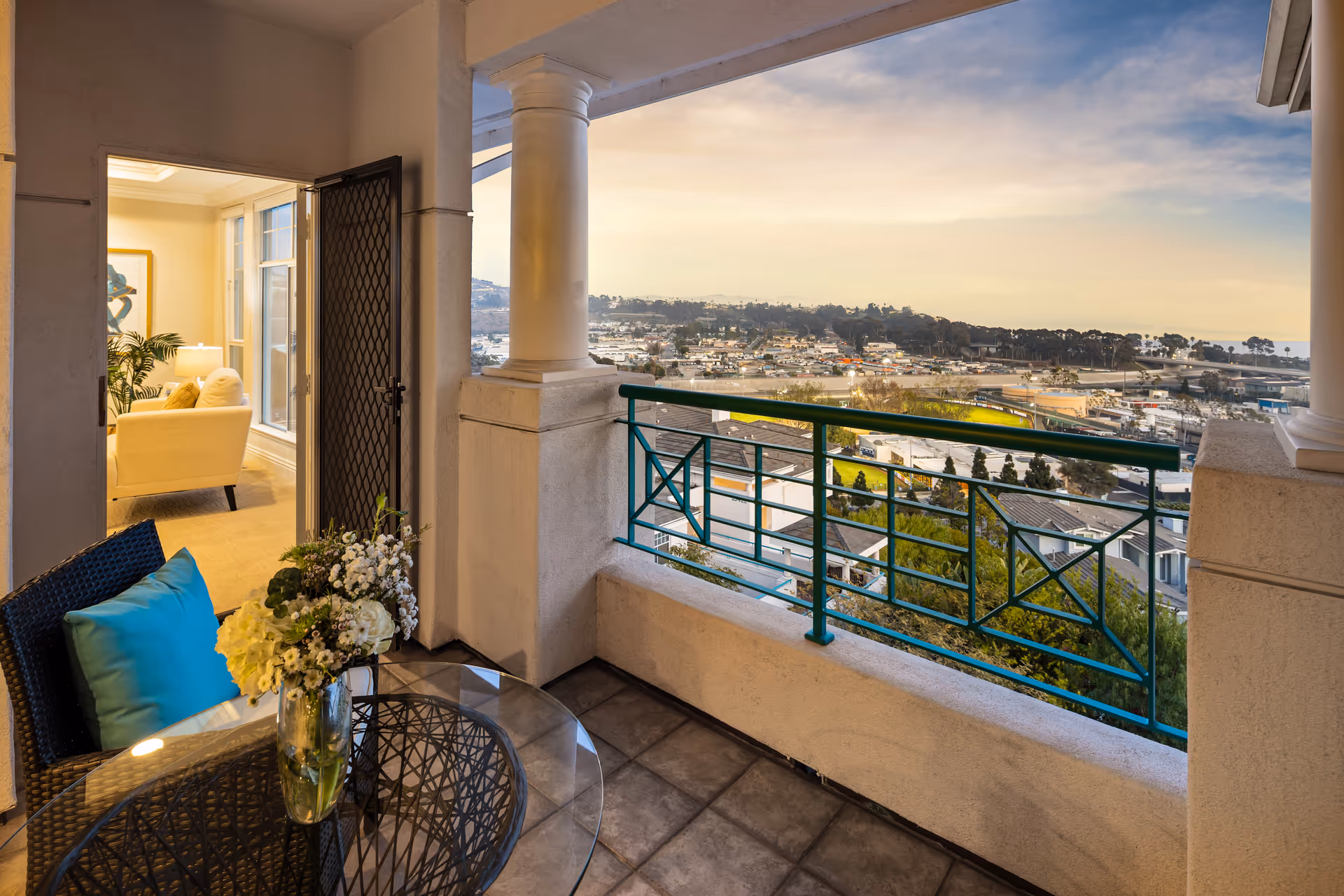 View from a balcony with a glass-top table and a wicker chair with a blue cushion. A vase with white flowers is on the table. The balcony has a decorative green railing and white columns. Through an open door, a cozy living room with a white armchair, a lamp, and a framed artwork is visible. The background shows a scenic view of a town and hills under a partly cloudy sky.