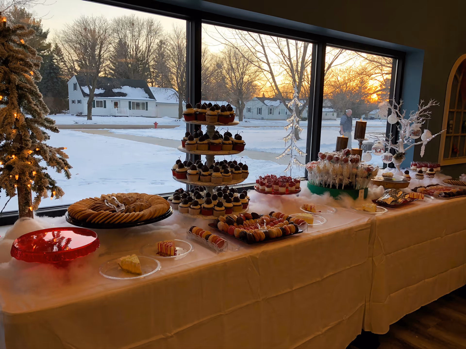 A dessert buffet table set up indoors near large windows showing a snowy outdoor scene at sunset. The table is covered with a white tablecloth and displays an assortment of desserts including cupcakes on a tiered stand, cookies, macarons, cake slices, and other sweet treats. A small decorated Christmas tree with lights and a white decorative tree with hanging ornaments are also on the table.